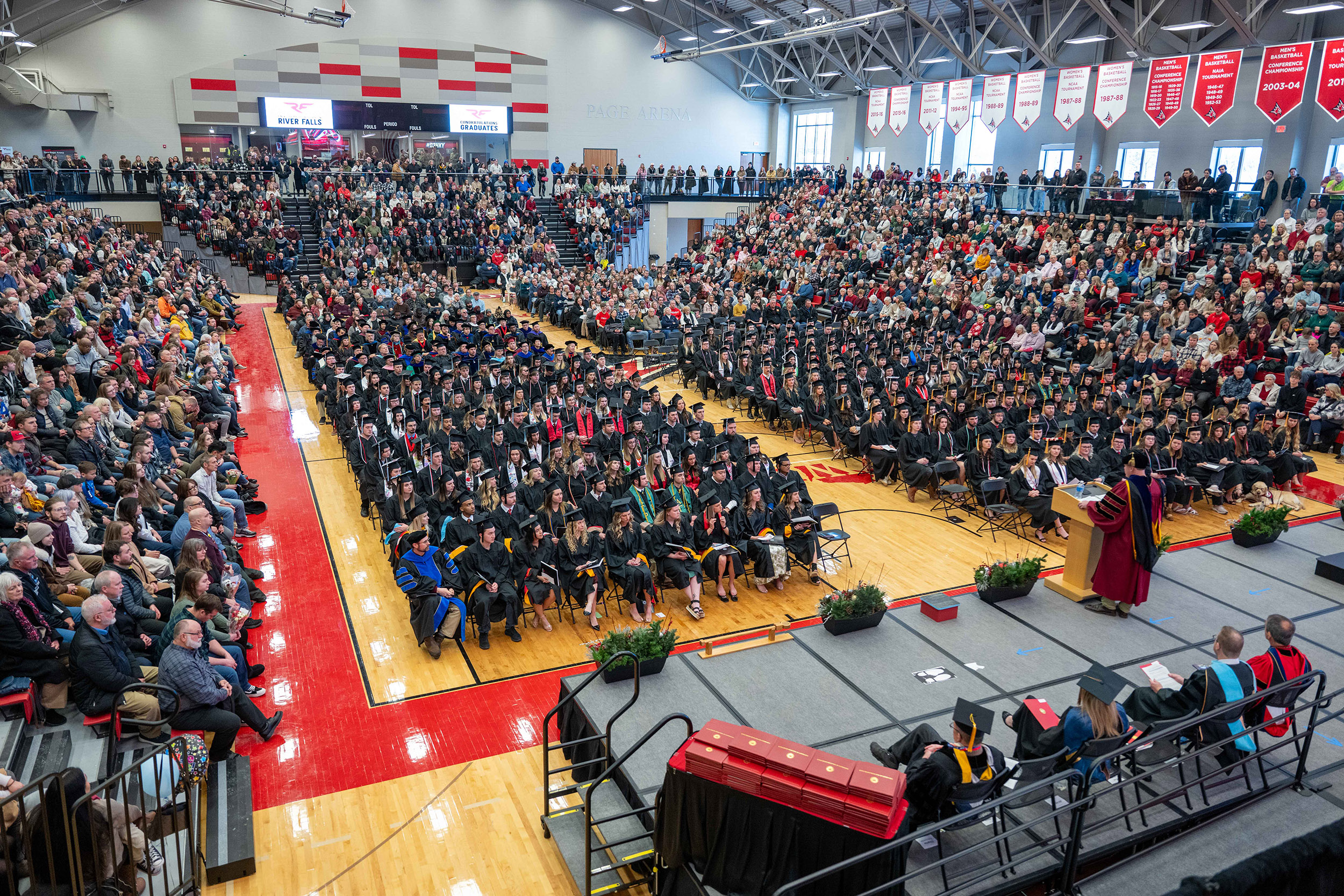 a gymnasium full of graduates wearing caps and gowns. The graduates are sitting in rows of chairs on the gym floor
