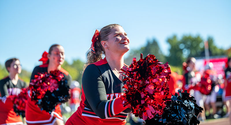 UWRF cheerleaders, wearing black and red uniforms and holding black and red pompoms, cheer at a football game.