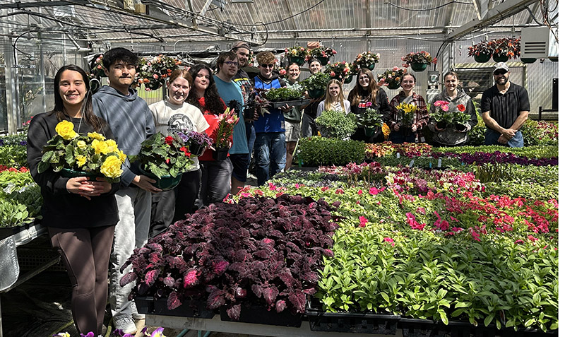 students stand in a greenhouse filled with plants while each holding hanging baskets filled with flowers