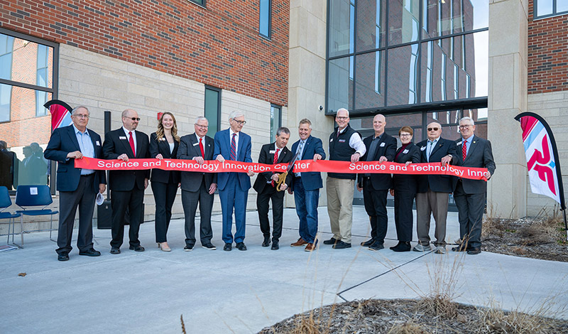 12 people stand in front of the new SciTech building holding a red ribbon while one cuts the ribbon with an oversized pair of scissors