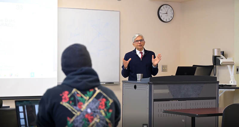 a male professor stands at a desk and speaks to students
