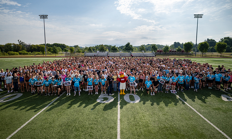 a large group of approximately 1300 new students stand on the football field at UWRF. THe Freddy Falcon mascot is at the front center of the photo.