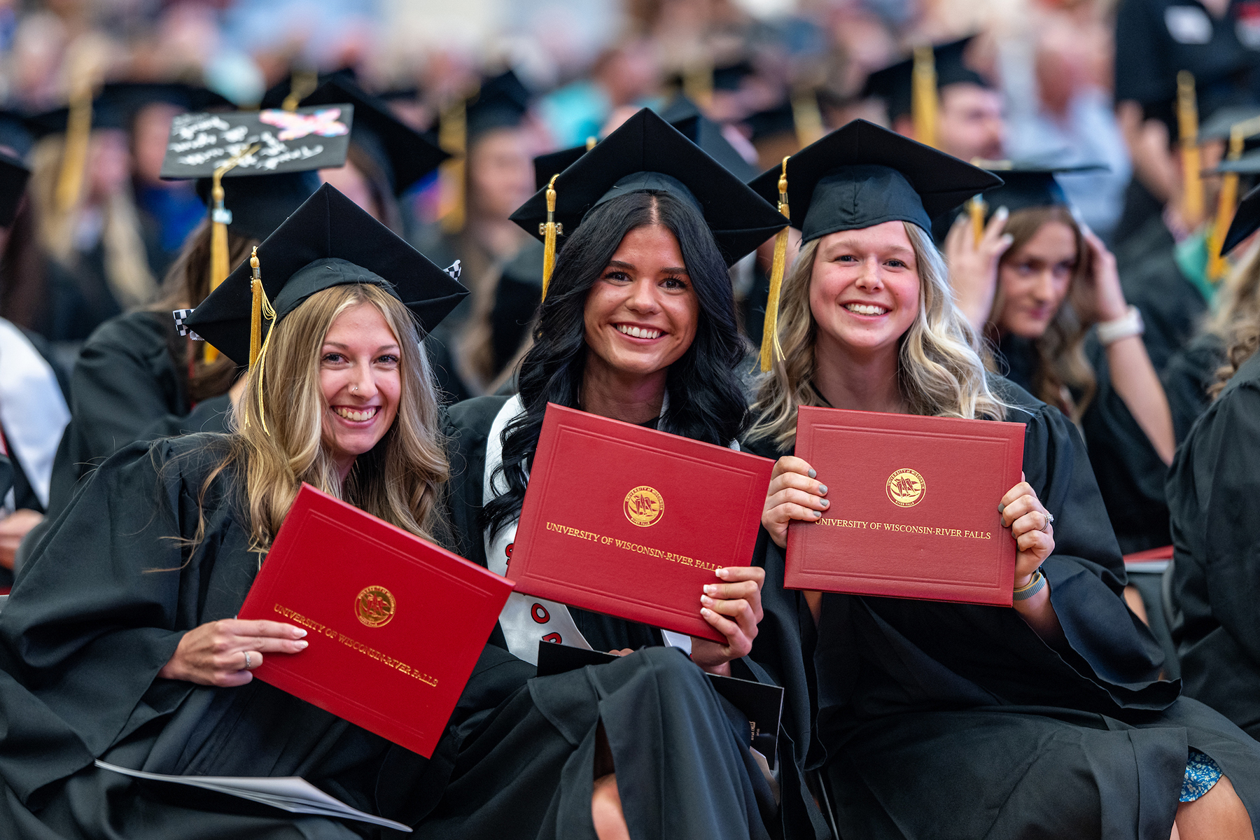 Three graduates in black caps and gowns hold red University of Wisconsin–River Falls diploma covers.