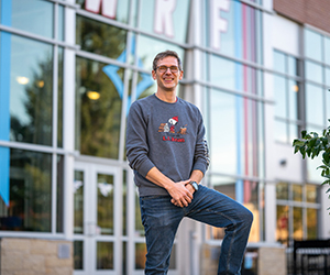 Man in a gray sweatshirt and jeans standing in front on the University Center entrance