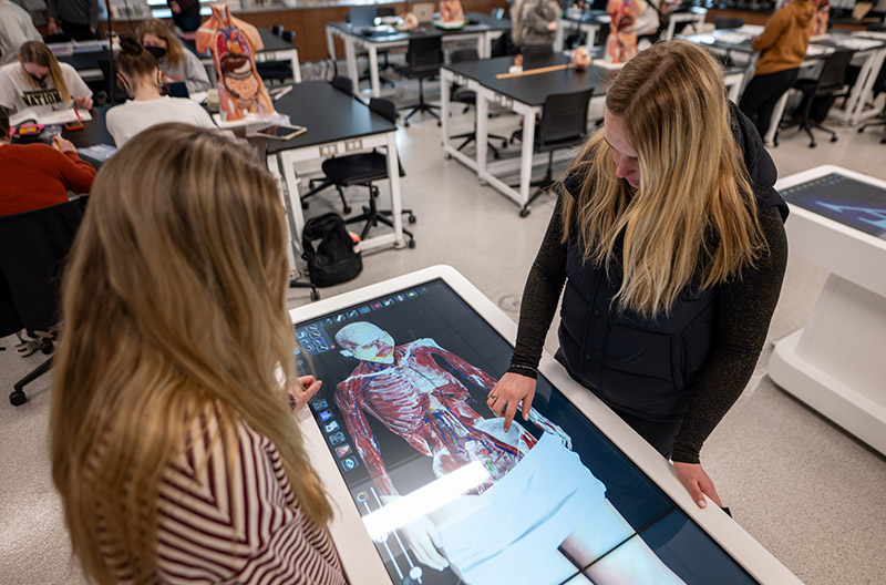 two students with long blonde hair stand over a virtual dissection table in a science lab. An image of a human cadaver is projected on the table.