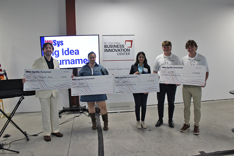 five people stand while holding oversized checks. The sign in the background says WiSys Big Idea Tournament